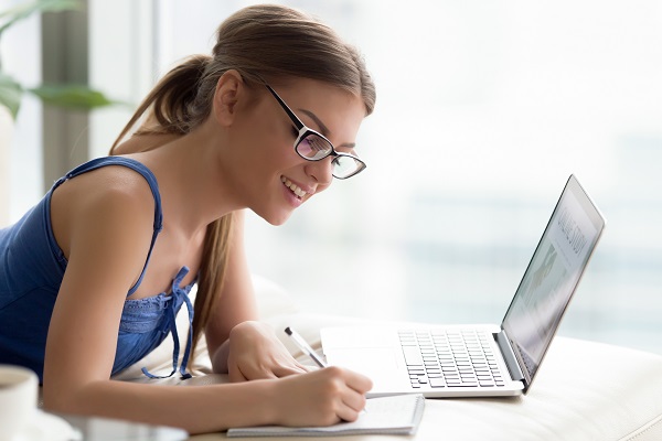 Smiling young woman studying online on laptop, making notes, sel